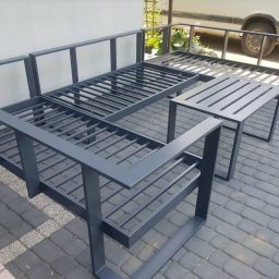 Roman Ko - Metalowy szkielet mebli ogrodowych w minimalistycznym stylu, z bliska na tle kostki brukowej i fragmentu budynku. Widoczne spawane łączenia i grafitowa kolorystyka.