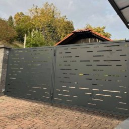 Roman Ko - Nowoczesna, grafitowa brama wjazdowa z poziomymi, prostokątnymi wycięciami, osadzona na kamiennych słupkach. Brukowana podstawa, w tle zieleń i fragment dachu.