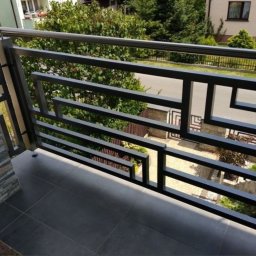 Roman Ko - Czarna, geometryczna balustrada balkonowa z elementami ze stali nierdzewnej, widok z góry na szare płytki balkonu i otoczenie budynku.