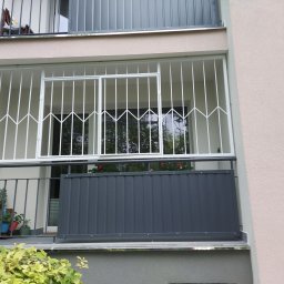 Roman Ko - Balkon z białą, metalową kratą zabezpieczającą okno i szarą, panelową balustradą. Na balkonie widoczne kwiaty w doniczkach.