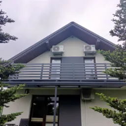 Szara, nowoczesna balustrada balkonowa z poziomymi szczeblami, zamontowana na domu z klimatyzatorami, otoczona zielonymi drzewami iglastymi.