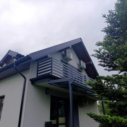 Roman Ko - Nowoczesny balkon z czarną, poziomą balustradą i szklanym zadaszeniem, widok z dołu na tle pochmurnego nieba i zielonego drzewa.