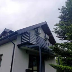 Nowoczesny balkon z czarną, poziomą balustradą i szklanym zadaszeniem, widok z dołu na tle pochmurnego nieba i zielonego drzewa.