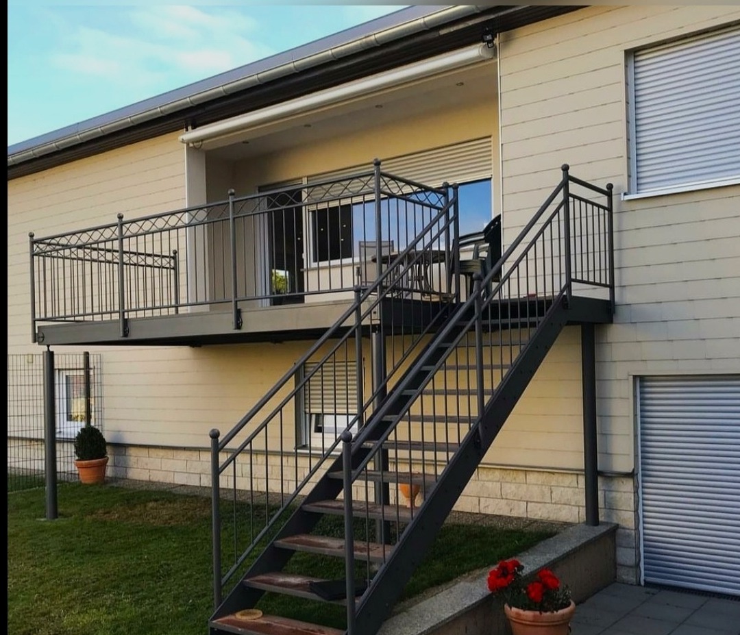 Zewnętrzne schody z metalową balustradą prowadzące na balkon z ozdobną, kutą barierką. Jasna elewacja budynku z roletami okiennymi i markizą.