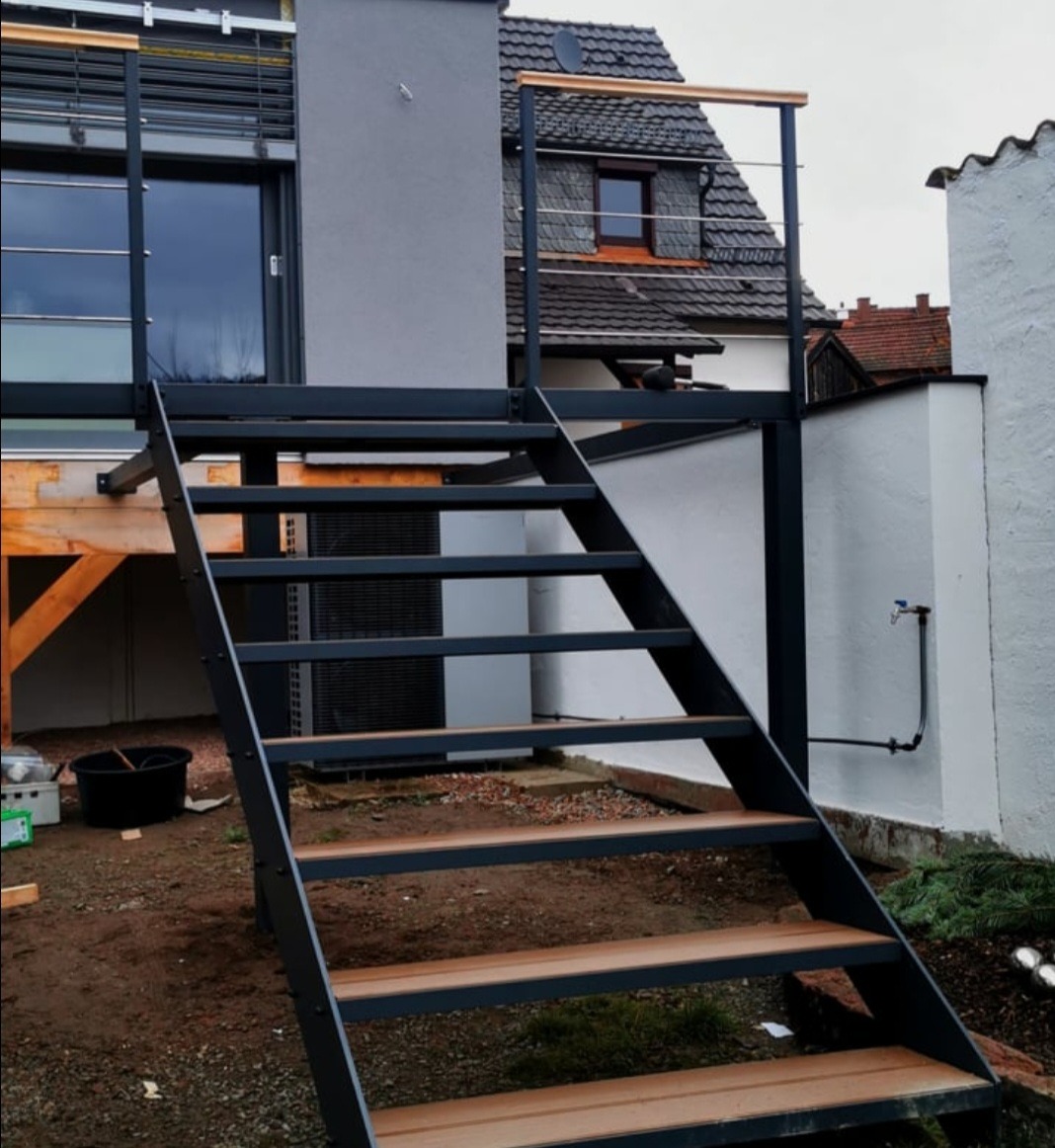 Stalowe schody z drewnianymi stopniami prowadzące na balkon z drewnianą poręczą. Konstrukcja przy budynku mieszkalnym, widok z dołu.