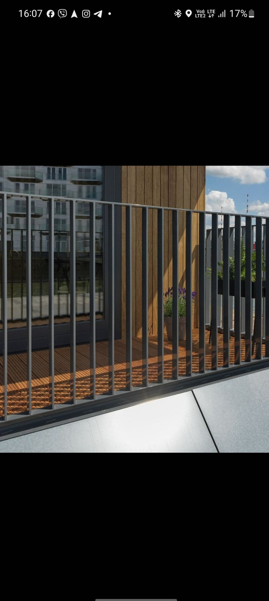 Nowoczesna balustrada balkonowa z pionowymi, szarymi szczeblami na tle drewnianej ściany i płytek. Widok na budynek w tle.