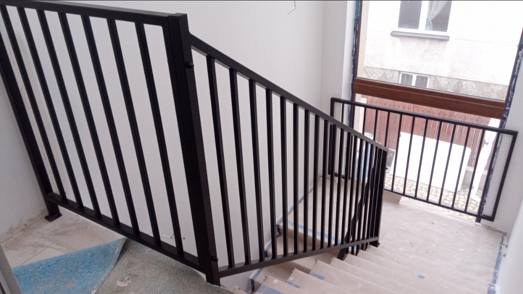 Nowoczesna, czarna balustrada schodowa z pionowymi szczebelkami, widok z góry na klatkę schodową w trakcie wykończenia, zabezpieczone schody, okno w tle.