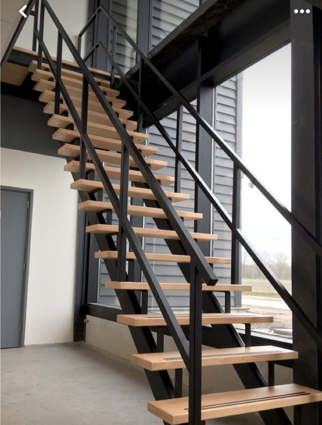 Nowoczesne schody wewnętrzne z drewnianymi stopniami i czarną, metalową konstrukcją, widoczne na tle okna z żaluzjami.