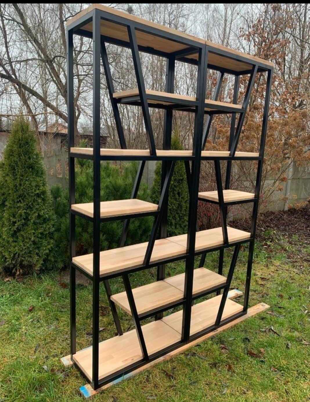 Metalowa konstrukcja regału z drewnianymi półkami, ustawiona na zewnątrz. Czarna rama, jasne drewno, nietypowy układ półek. Widok z bliska na tle zieleni i drzew.