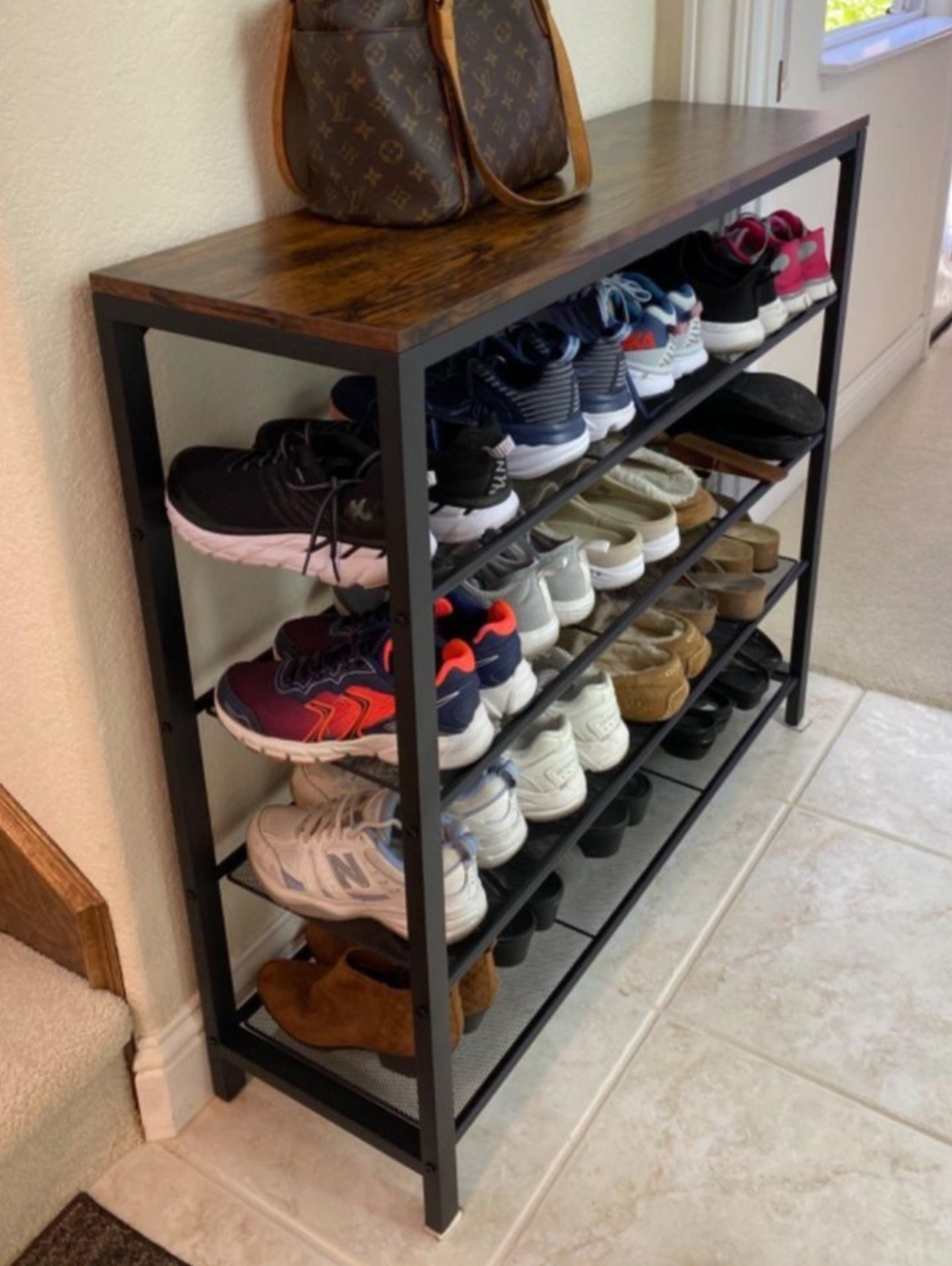 Metalowy regał na buty z drewnianym blatem, wypełniony różnorodnym obuwiem, obok schodów i torby. Minimalistyczny design, jasne płytki podłogowe.