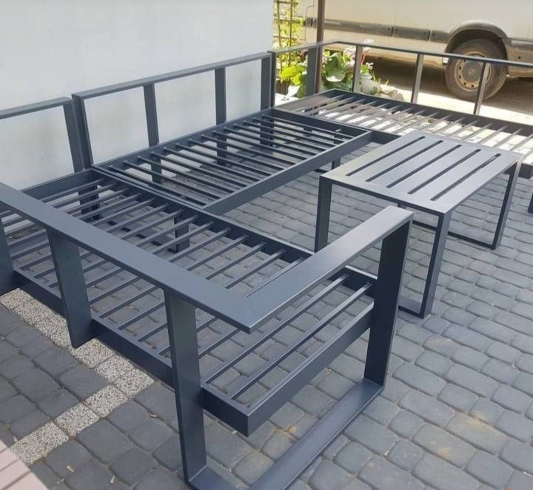 Metalowy szkielet mebli ogrodowych w minimalistycznym stylu, z bliska na tle kostki brukowej i fragmentu budynku. Widoczne spawane łączenia i grafitowa kolorystyka.