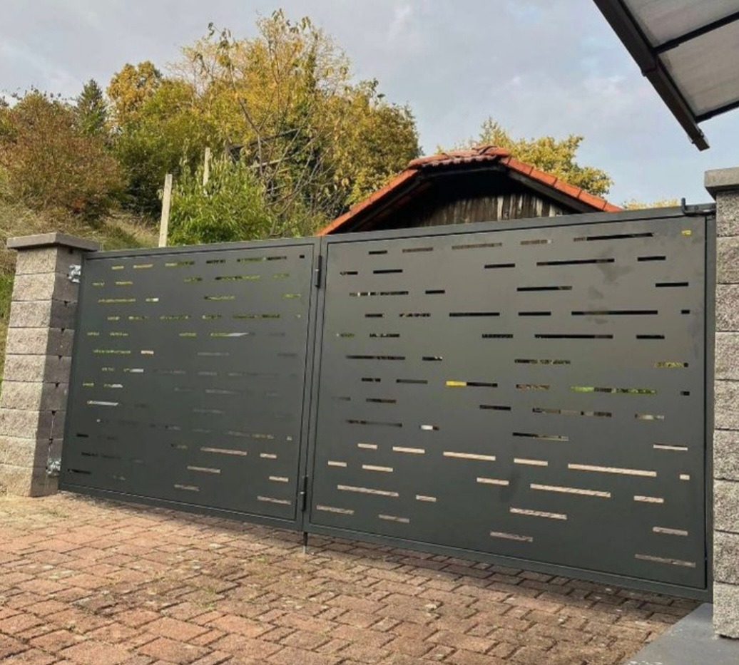 Nowoczesna, grafitowa brama wjazdowa z poziomymi, prostokątnymi wycięciami, osadzona na kamiennych słupkach. Brukowana podstawa, w tle zieleń i fragment dachu.