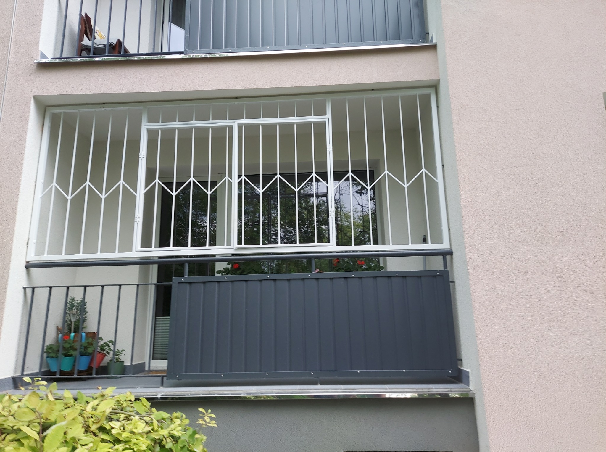 Balkon z białą, metalową kratą zabezpieczającą okno i szarą, panelową balustradą. Na balkonie widoczne kwiaty w doniczkach.