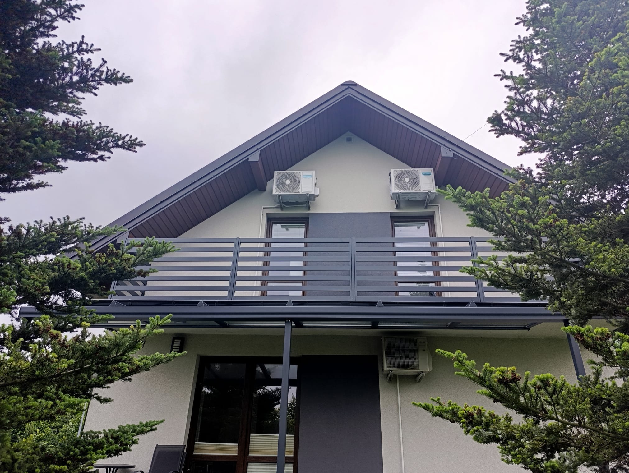 Szara, nowoczesna balustrada balkonowa z poziomymi szczeblami, zamontowana na domu z klimatyzatorami, otoczona zielonymi drzewami iglastymi.