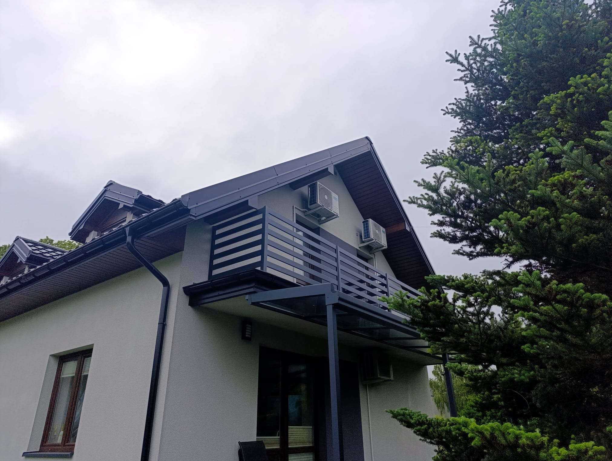 Nowoczesny balkon z czarną, poziomą balustradą i szklanym zadaszeniem, widok z dołu na tle pochmurnego nieba i zielonego drzewa.