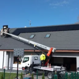 Montaż paneli słonecznych na dachu domu jednorodzinnego z użyciem podnośnika koszowego; pracownik w żółtej kamizelce na platformie. Błękitne niebo w tle.