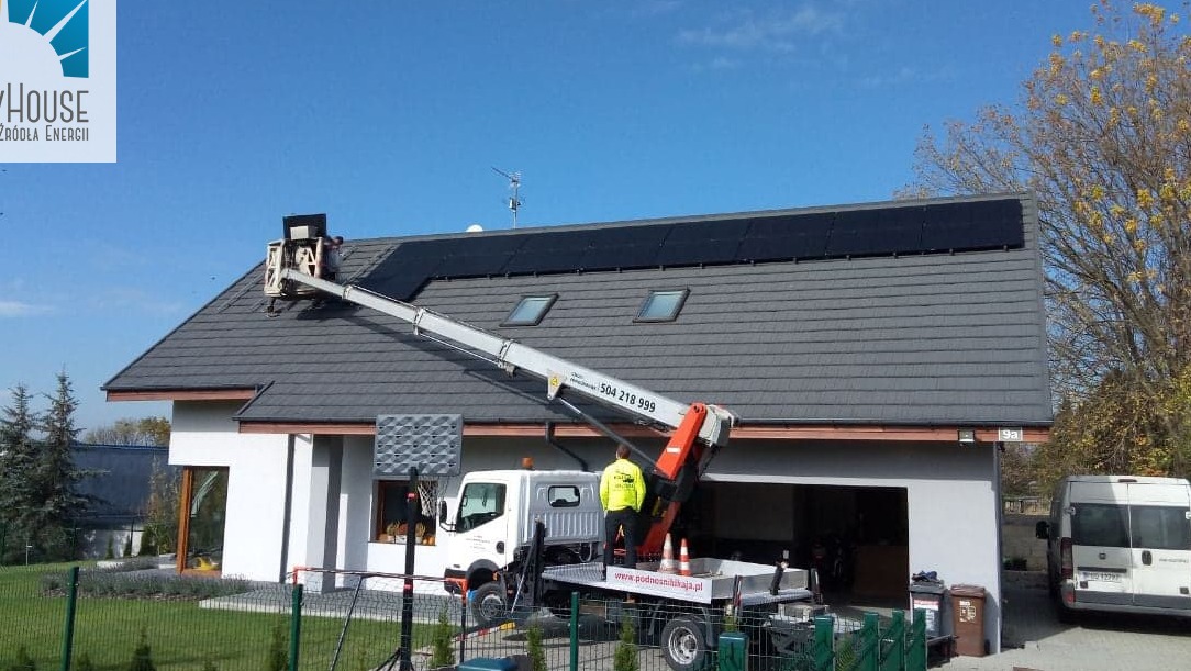 Montaż paneli słonecznych na dachu domu jednorodzinnego z użyciem podnośnika koszowego; pracownik w żółtej kamizelce na platformie. Błękitne niebo w tle.
