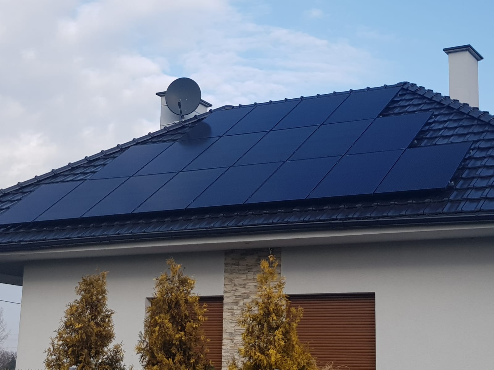 Czarny panel fotowoltaiczny na dachu domu z brązową dachówką i anteną satelitarną. Na pierwszym planie drzewa iglaste i fragment fasady z roletami.