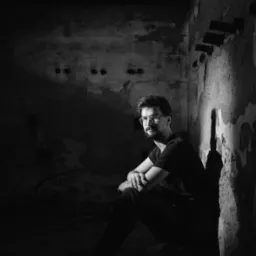 Czarno-biały portret mężczyzny w okularach, siedzącego w rogu zniszczonego pomieszczenia, oświetlonego kontrastowym światłem, z widoczną fakturą ściany.