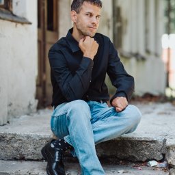 Marcin Szumański - Mężczyzna w czarnej koszuli i jeansach siedzi na betonowych schodach, podpierając brodę dłonią, zamyślony wzrok skierowany w stronę widza. W tle budynek z oknami.