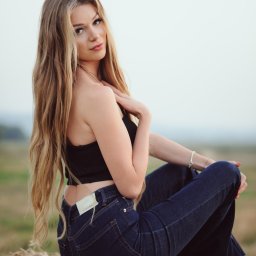 Marcin Szumański - Blondwłosa kobieta w czarnym topie i dżinsach siedzi na słomianej beli na tle rozmytego krajobrazu. Uwagę przykuwa jej spojrzenie i naturalność scenerii.