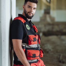 Marcin Szumański - Sesja biznesowa ratownika medycznego w kamizelce i spodniach w odcieniach czerwieni, oparty o ścianę w surowym wnętrzu. Ujęcie portretowe, profesjonalne.