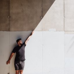 Marcin Szumański - Mężczyzna w czarnym polo i szarych szortach pozuje na tle betonowej ściany z wyraźnym cieniem, wyciągając rękę w górę. Ujęcie z dołu podkreśla geometrię ściany.