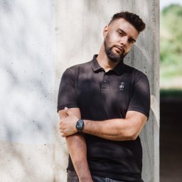 Marcin Szumański - Mężczyzna z brodą w czarnej koszulce polo opiera się o betonowy filar. Na nadgarstku ma smartwatcha. Ujęcie w plenerze, w tle zieleń.