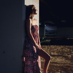 Marcin Szumański - Portret kobiety w kwiecistej sukience, oparta o betonowy filar w Dębicy. Ujęcie w kontraście światła i cienia, z widoczną fakturą podłoża i butami sportowymi.