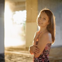 Marcin Szumański - Uśmiechnięta brunetka w sukience pozuje na tle surowego, betonowego wnętrza z promieniami słońca. Nietypowe oświetlenie i surowy kontrast dodają unikalnego charakteru.