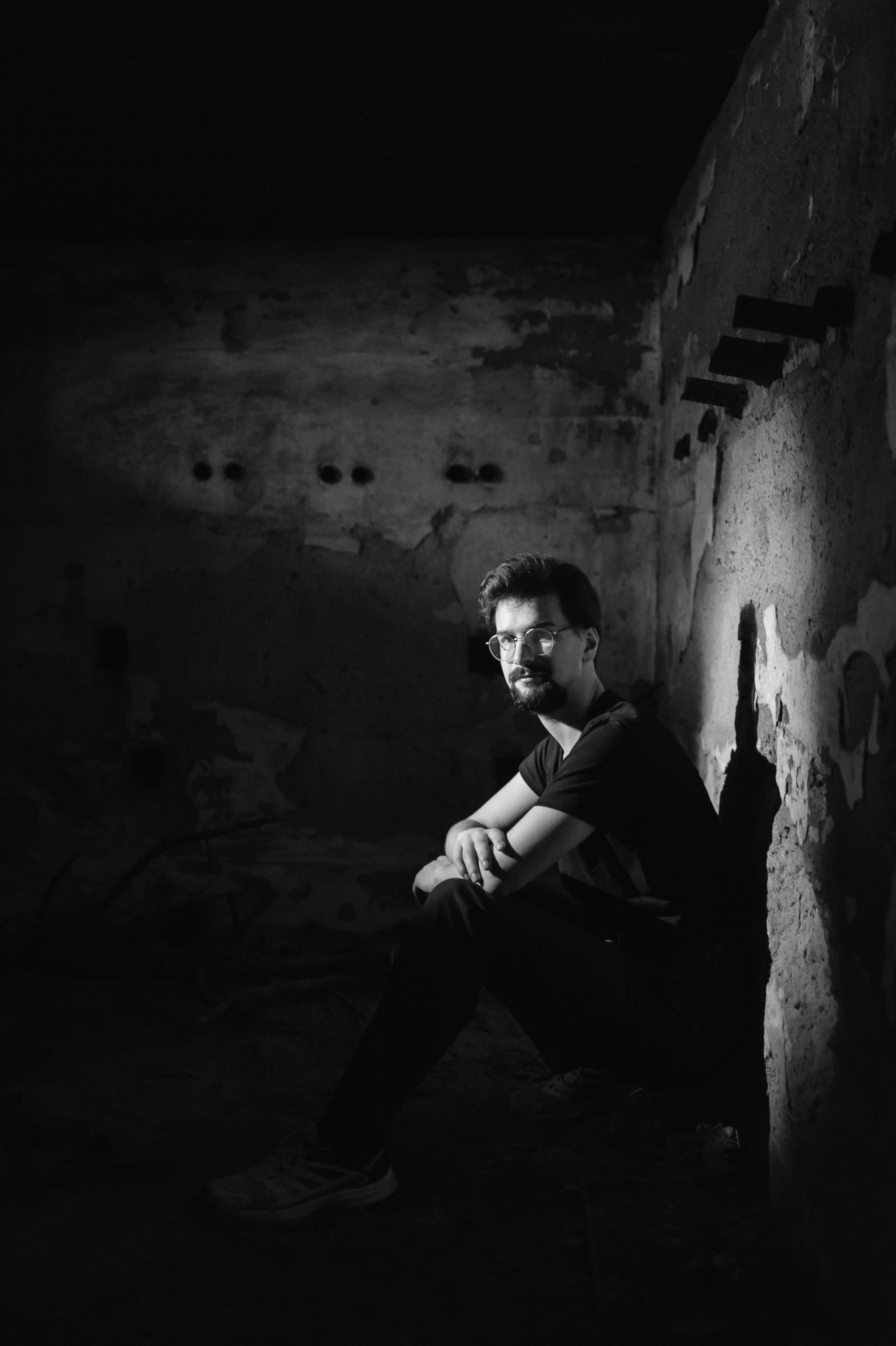 Czarno-biały portret mężczyzny w okularach, siedzącego w rogu zniszczonego pomieszczenia, oświetlonego kontrastowym światłem, z widoczną fakturą ściany.