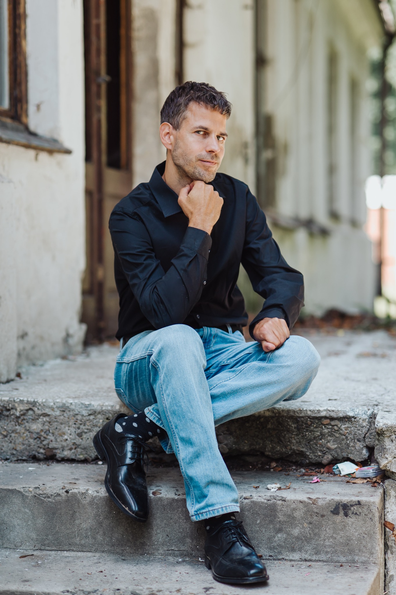 Mężczyzna w czarnej koszuli i jeansach siedzi na betonowych schodach, podpierając brodę dłonią, zamyślony wzrok skierowany w stronę widza. W tle budynek z oknami.