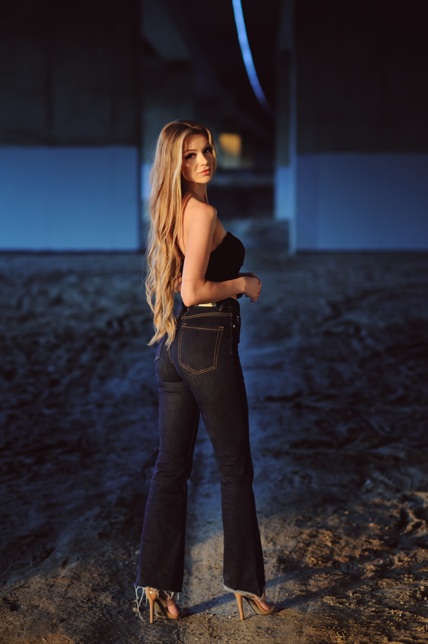 Portret kobiety z długimi blond włosami, ubrana w czarny top i dżinsy, pozuje patrząc przez ramię w plenerze z oświetleniem podkreślającym jej sylwetkę.