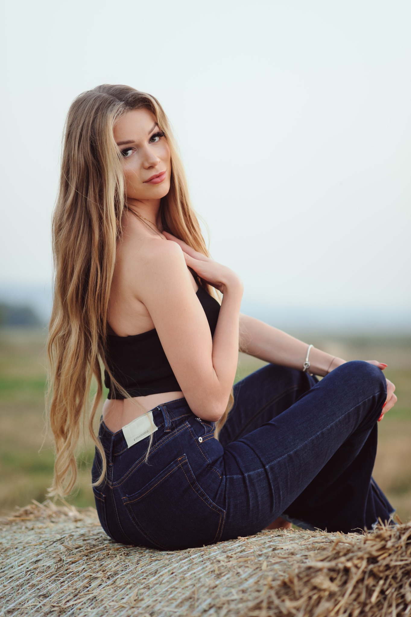 Blondwłosa kobieta w czarnym topie i dżinsach siedzi na słomianej beli na tle rozmytego krajobrazu. Uwagę przykuwa jej spojrzenie i naturalność scenerii.