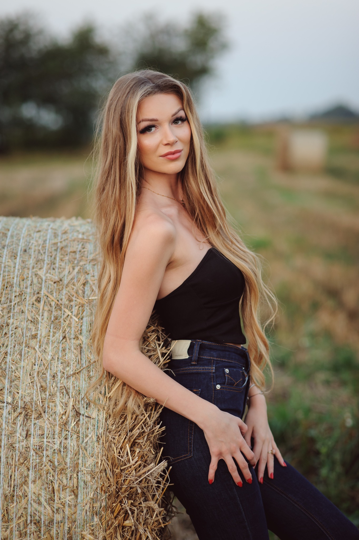 Portret kobiety z długimi blond włosami, opierającej się o belę siana na tle pola. Ubrana w czarny top i ciemne jeansy. Delikatny makijaż.
