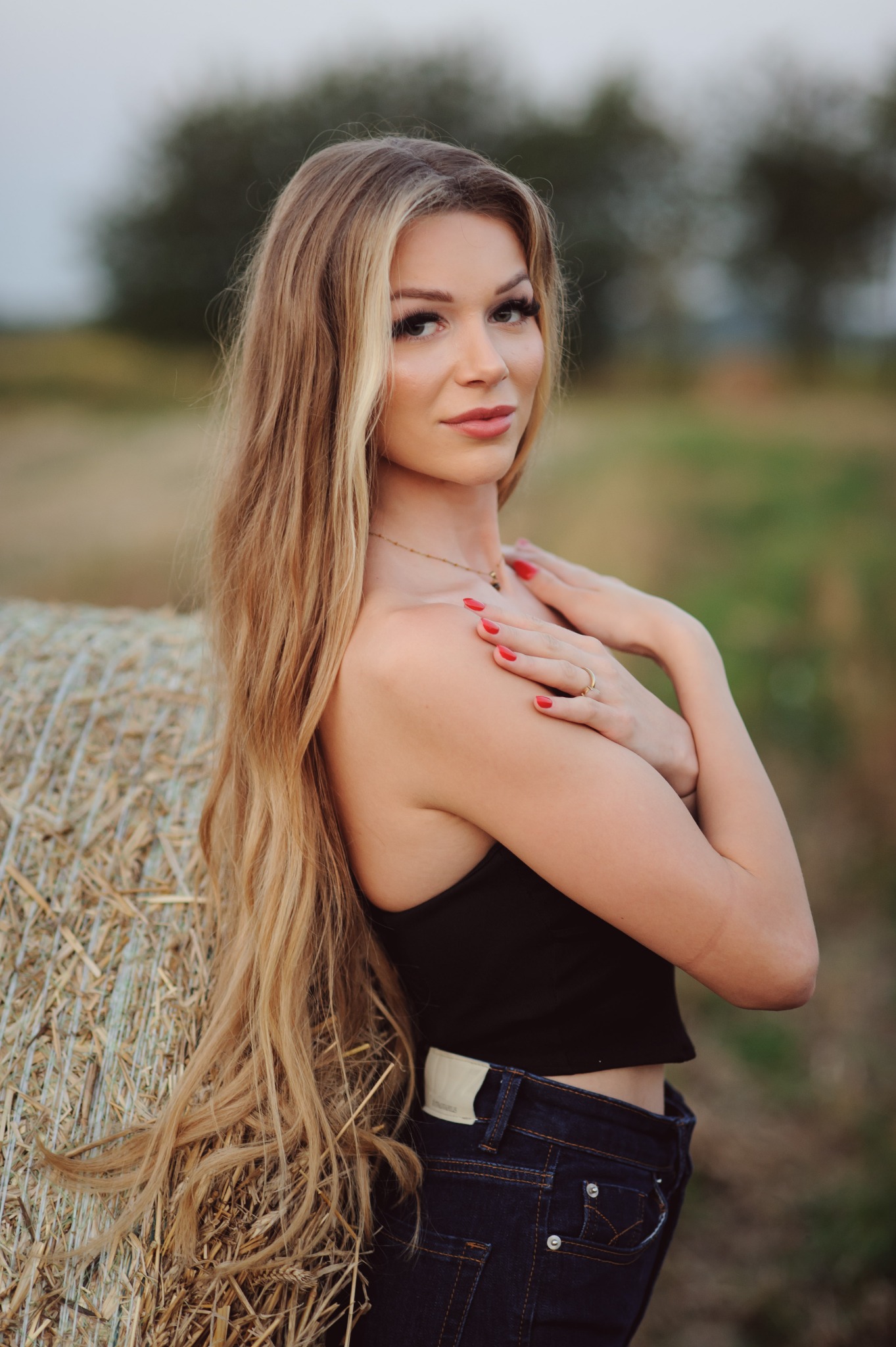 Portret kobiety o długich blond włosach, opierającej się o belę siana na polu, z delikatnym makijażem i czerwoną emalią na paznokciach, w czarnym topie i dżinsach.