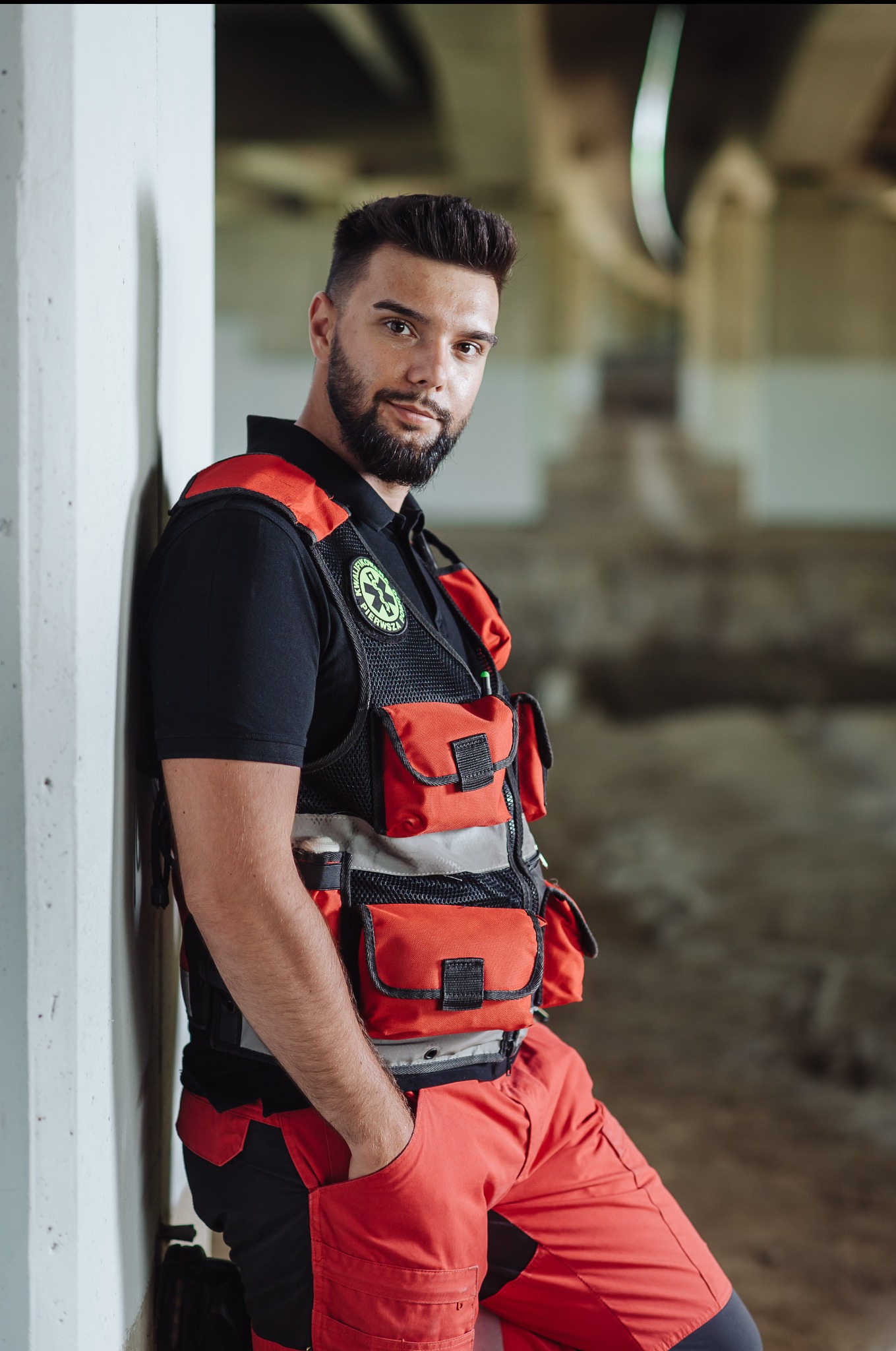 Sesja biznesowa ratownika medycznego w kamizelce i spodniach w odcieniach czerwieni, oparty o ścianę w surowym wnętrzu. Ujęcie portretowe, profesjonalne.