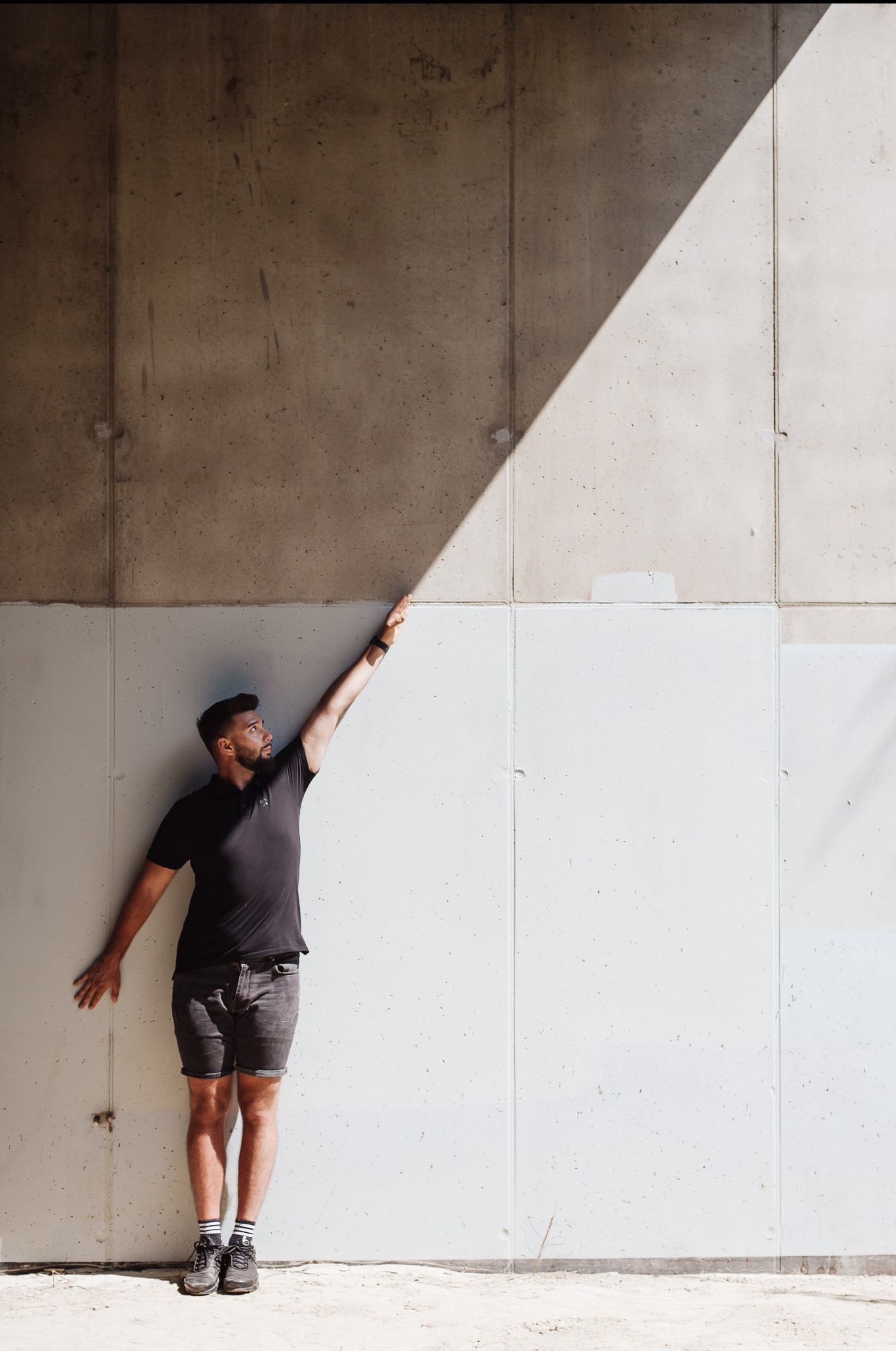 Mężczyzna w czarnym polo i szarych szortach pozuje na tle betonowej ściany z wyraźnym cieniem, wyciągając rękę w górę. Ujęcie z dołu podkreśla geometrię ściany.