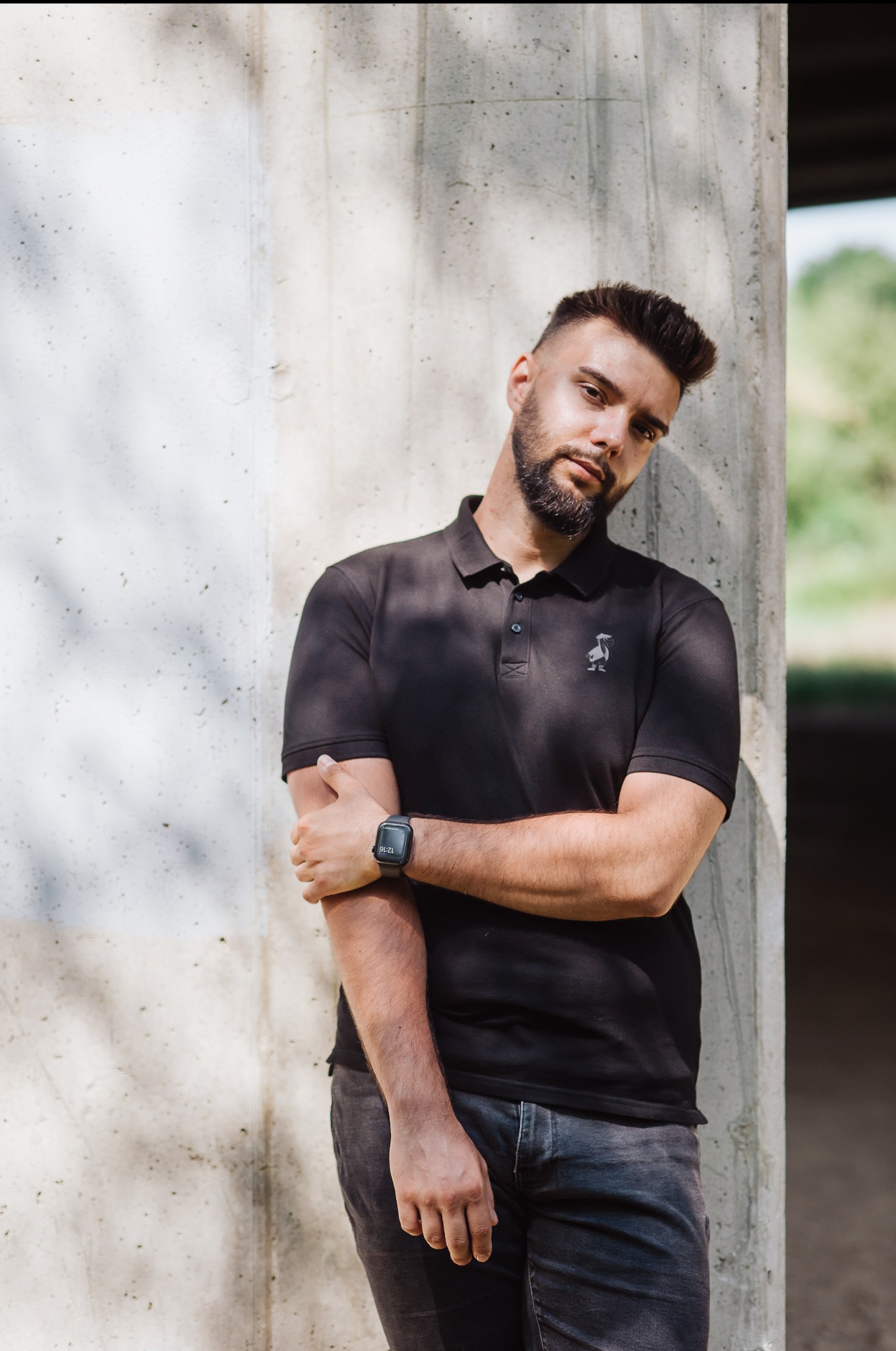 Mężczyzna z brodą w czarnej koszulce polo opiera się o betonowy filar. Na nadgarstku ma smartwatcha. Ujęcie w plenerze, w tle zieleń.