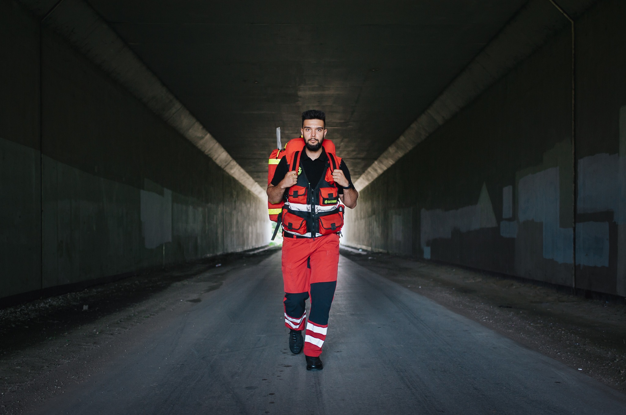 Ratownik medyczny w czerwonym uniformie i z plecakiem idzie tunelem, patrząc wprost. Ujęcie centralne, oświetlenie tunelu podkreśla postać.