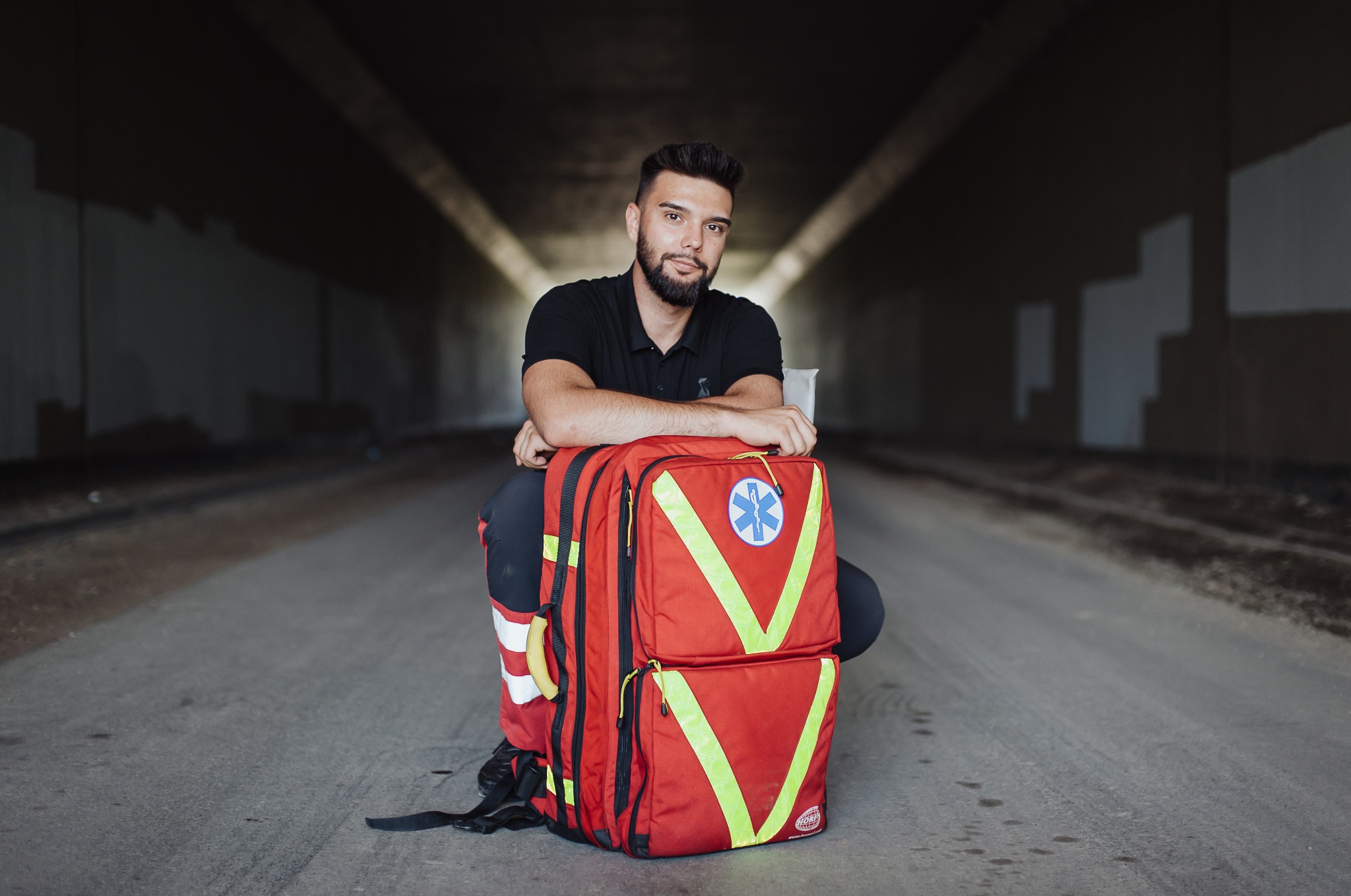 Ratownik medyczny w kucki opiera się o czerwony plecak medyczny z symbolem Eskulapa na tle betonowego tunelu. Uśmiechnięty mężczyzna z brodą pozuje do zdjęcia.
