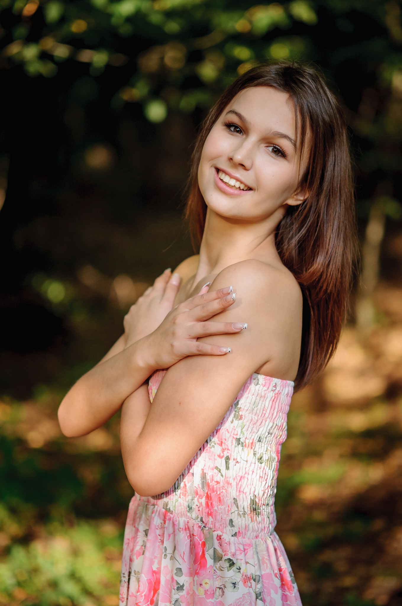 Uśmiechnięta brunetka w letniej sukience, z rękami skrzyżowanymi na ramionach, pozuje na tle zamazanej zieleni i brązów, prezentując manicure z białymi wzorami.