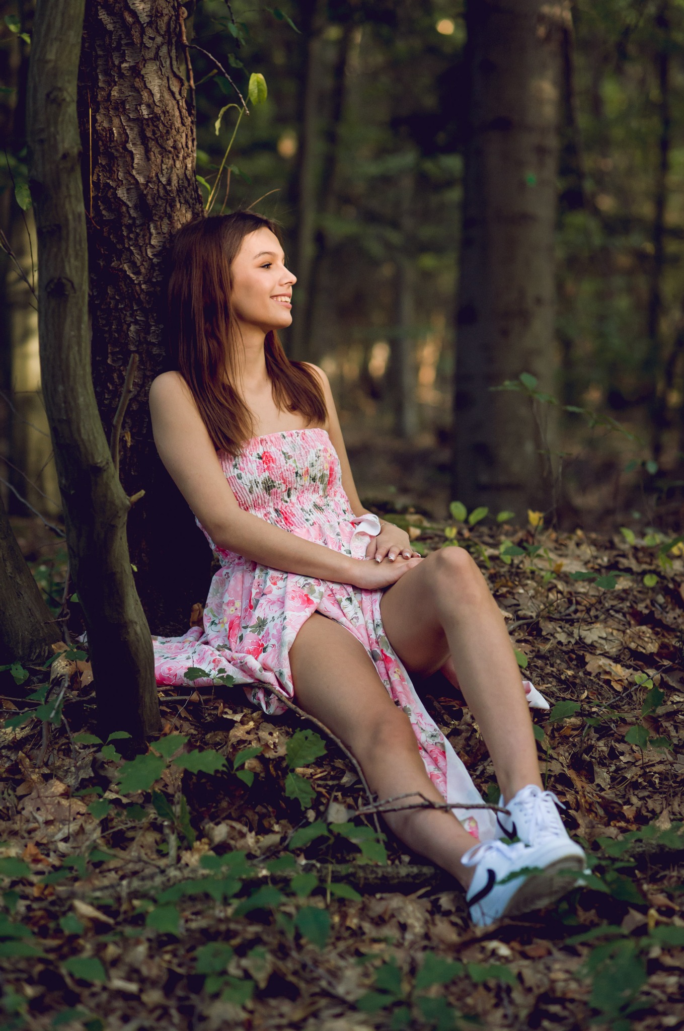 Uśmiechnięta brunetka w kwiecistej sukience i białych sneakersach opiera się o drzewo w lesie. Delikatne światło pada na jej twarz. Tło stanowią wysokie drzewa.