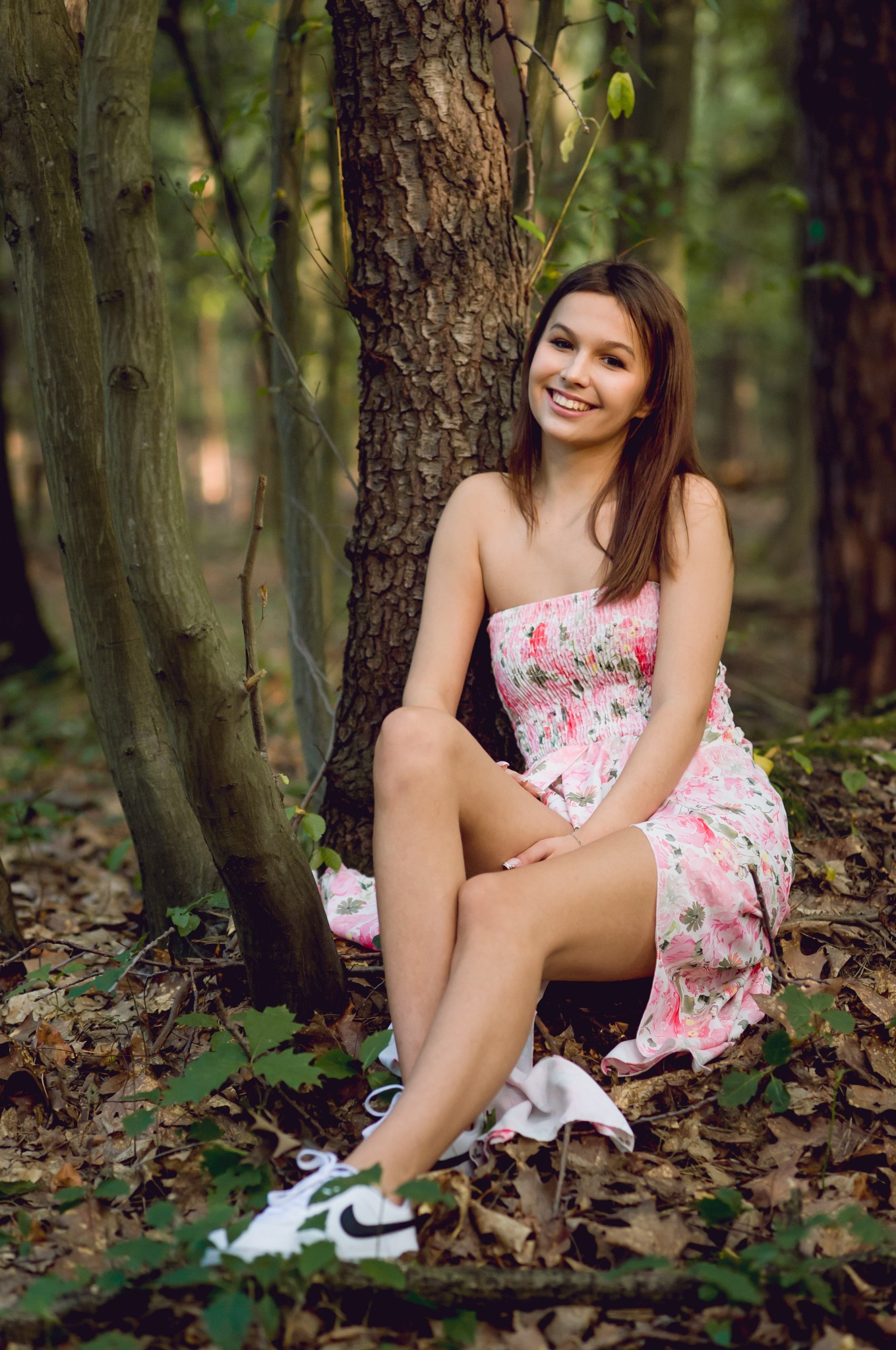 Uśmiechnięta brunetka w kwiecistej sukience siedzi oparta o drzewo w lesie. Białe sneakersy z czarnym logo. Naturalne światło, leśne otoczenie.