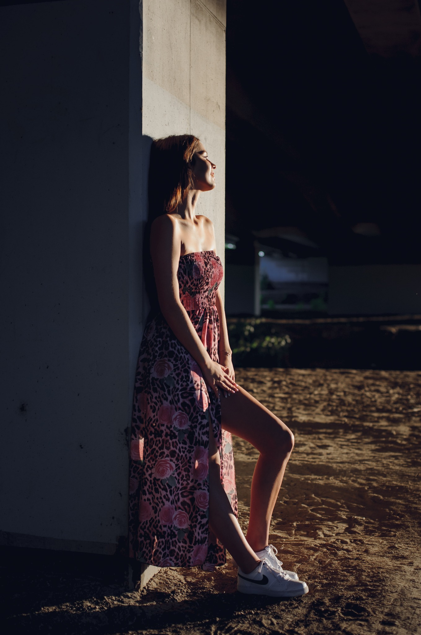 Portret kobiety w kwiecistej sukience, oparta o betonowy filar w Dębicy. Ujęcie w kontraście światła i cienia, z widoczną fakturą podłoża i butami sportowymi.