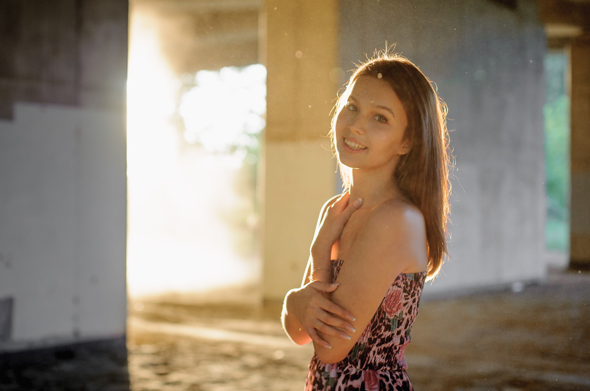 Uśmiechnięta brunetka w sukience pozuje na tle surowego, betonowego wnętrza z promieniami słońca. Nietypowe oświetlenie i surowy kontrast dodają unikalnego charakteru.