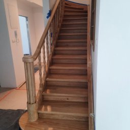 Juliusz Jacek Nowierski - Jasne, drewniane schody z balustradą w trakcie montażu, zabezpieczone niebieską taśmą malarską. Widok z dołu schodów na białe ściany i oświetlenie.