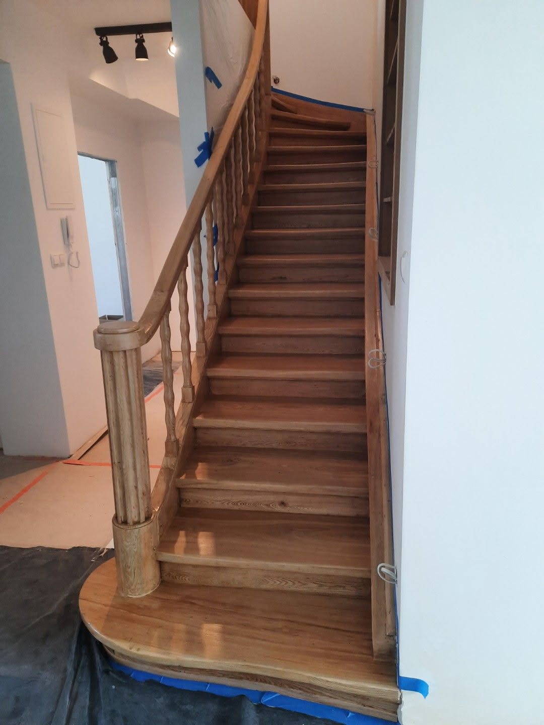 Jasne, drewniane schody z balustradą w trakcie montażu, zabezpieczone niebieską taśmą malarską. Widok z dołu schodów na białe ściany i oświetlenie.