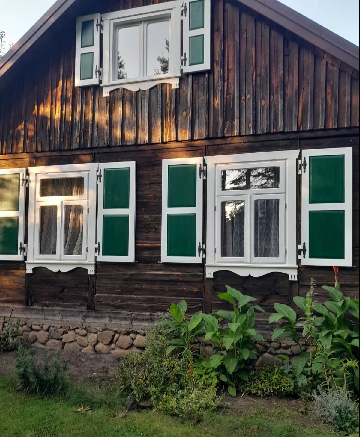 Drewniany dom z zielonymi okiennicami, oświetlony ciepłym światłem zachodzącego słońca, otoczony zielenią. Widoczne detale drewnianej elewacji i białe ramy okien.