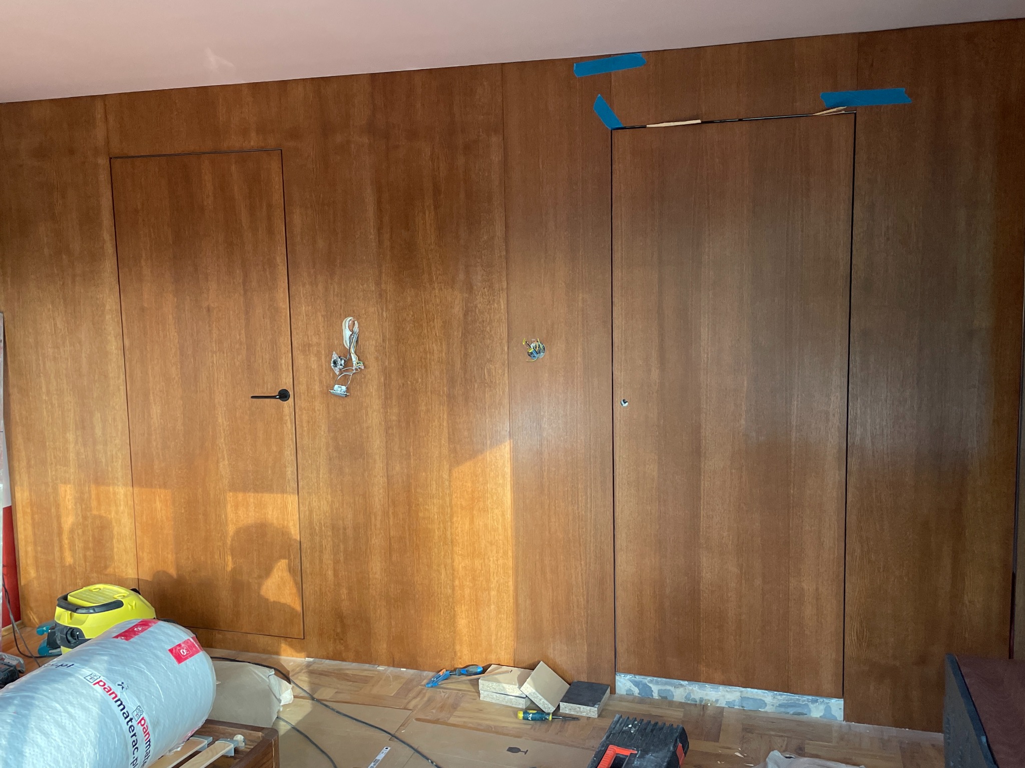 Montaż paneli drewnianych z ukrytymi drzwiami w trakcie prac wykończeniowych. Widoczne narzędzia, materiały izolacyjne i taśma malarska.